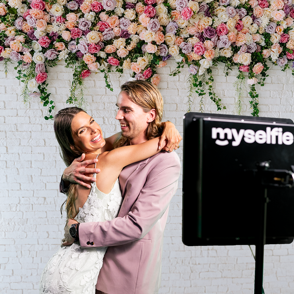 Brudepar krammer foran en blomstervæg ved siden af Myselfie photo booth til bryllup
