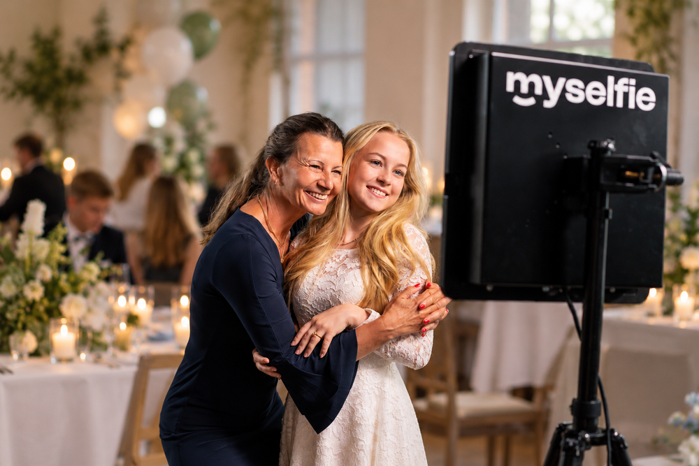 Konfirmand i hvid kjole krammer sin mor foran Myselfie photo booth til konfirmationsfesten