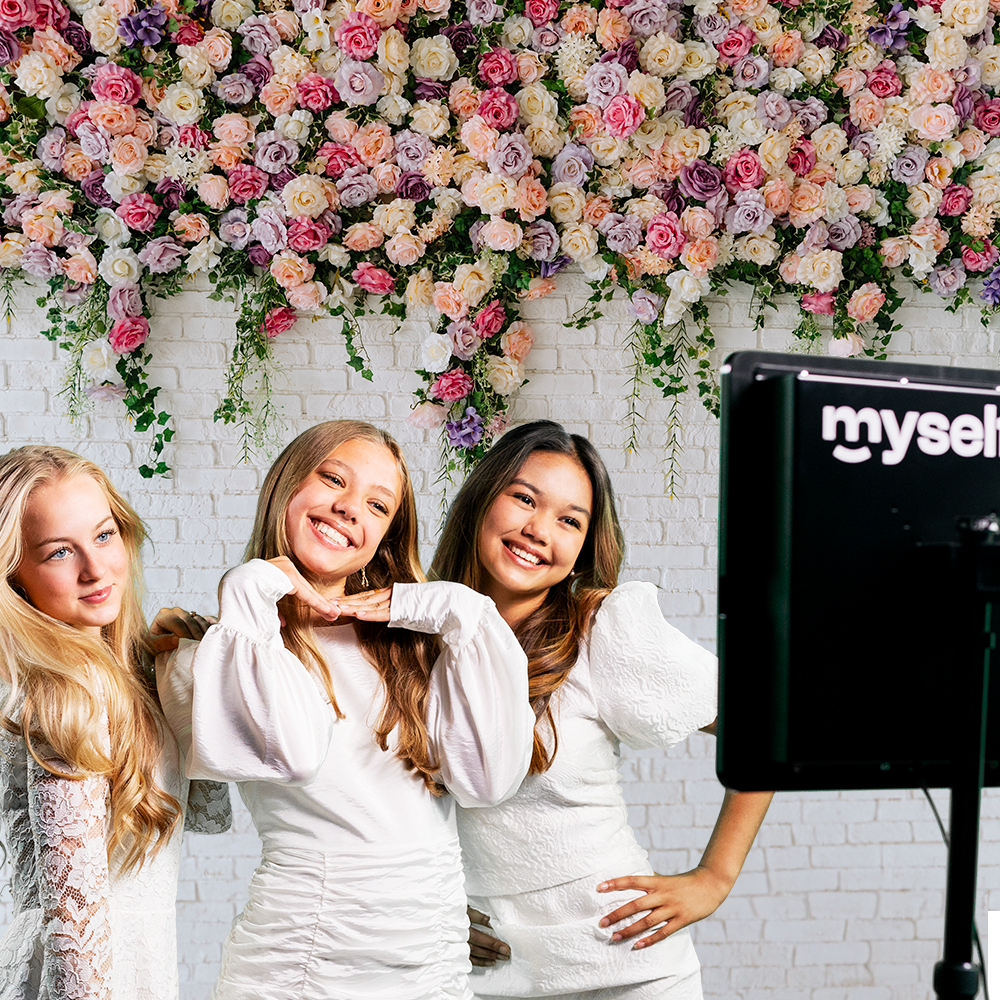 Konfirmand og veninder i hvidt griner foran Myselfie photo booth og en blomstervæg
